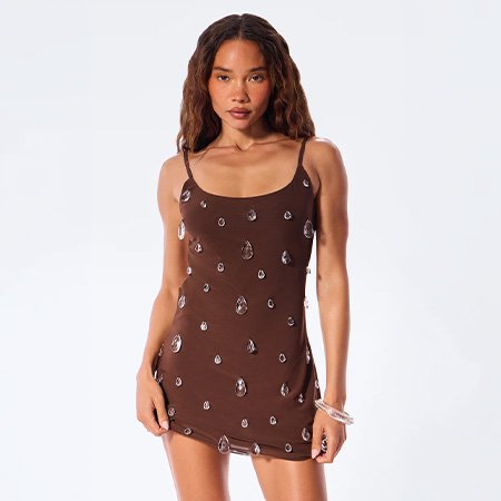 Woman wearing an embellished brown mini dress with crystal teardrops from a new fashion collection, posing against a white studio background.