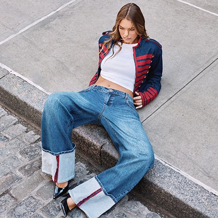 Woman wearing wide-leg cuffed jeans and a blue and red military jacket sits on a curb, showcasing trendy fall street style fashion.