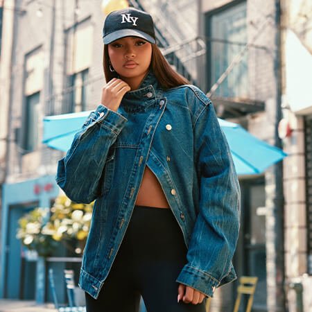 Woman in an oversized blue denim jacket, NY baseball cap, and black leggings poses on a city street, showcasing trendy urban street style.